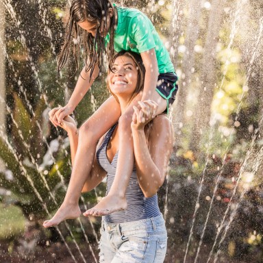 photo of a woman with a little girl on her shoulders, playing in a backyard sprinkler